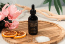 Bottle of 'Velvet Drops' luxury warmer oil on a wooden tray with flowers and oranges.