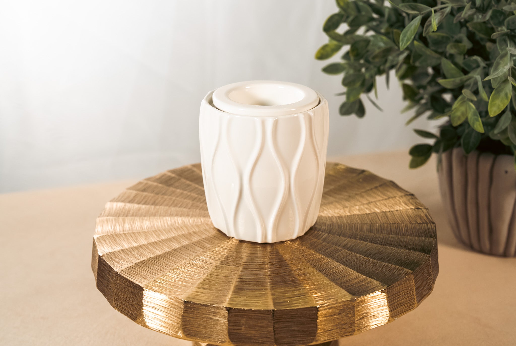 A textured, white fragrance warmer on a gold pedestal with a plant in the background