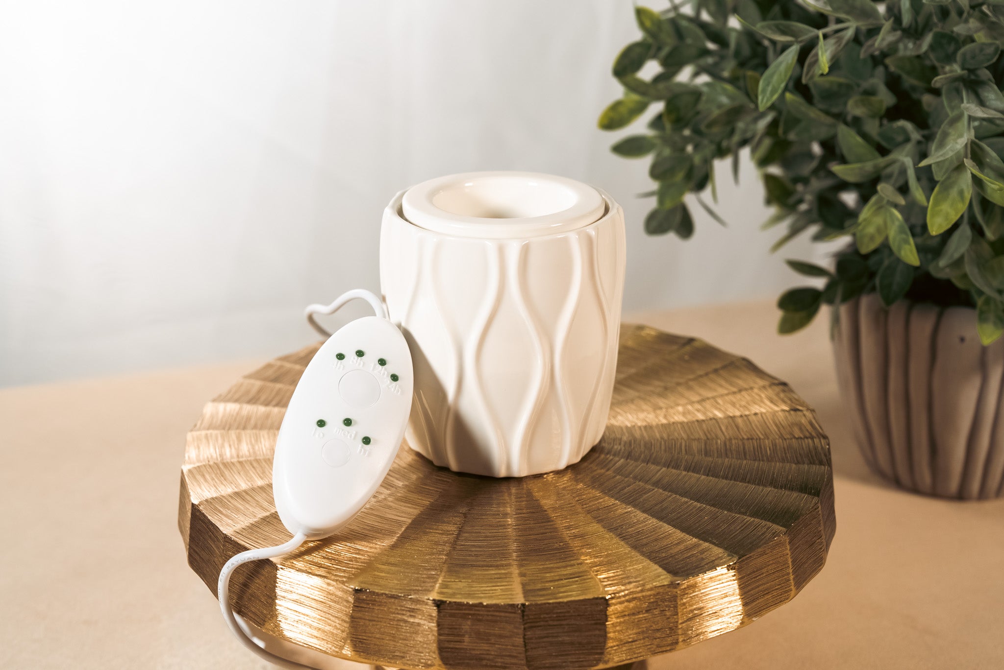 A textured, white fragrance warmer on a gold pedestal with a plant in the background