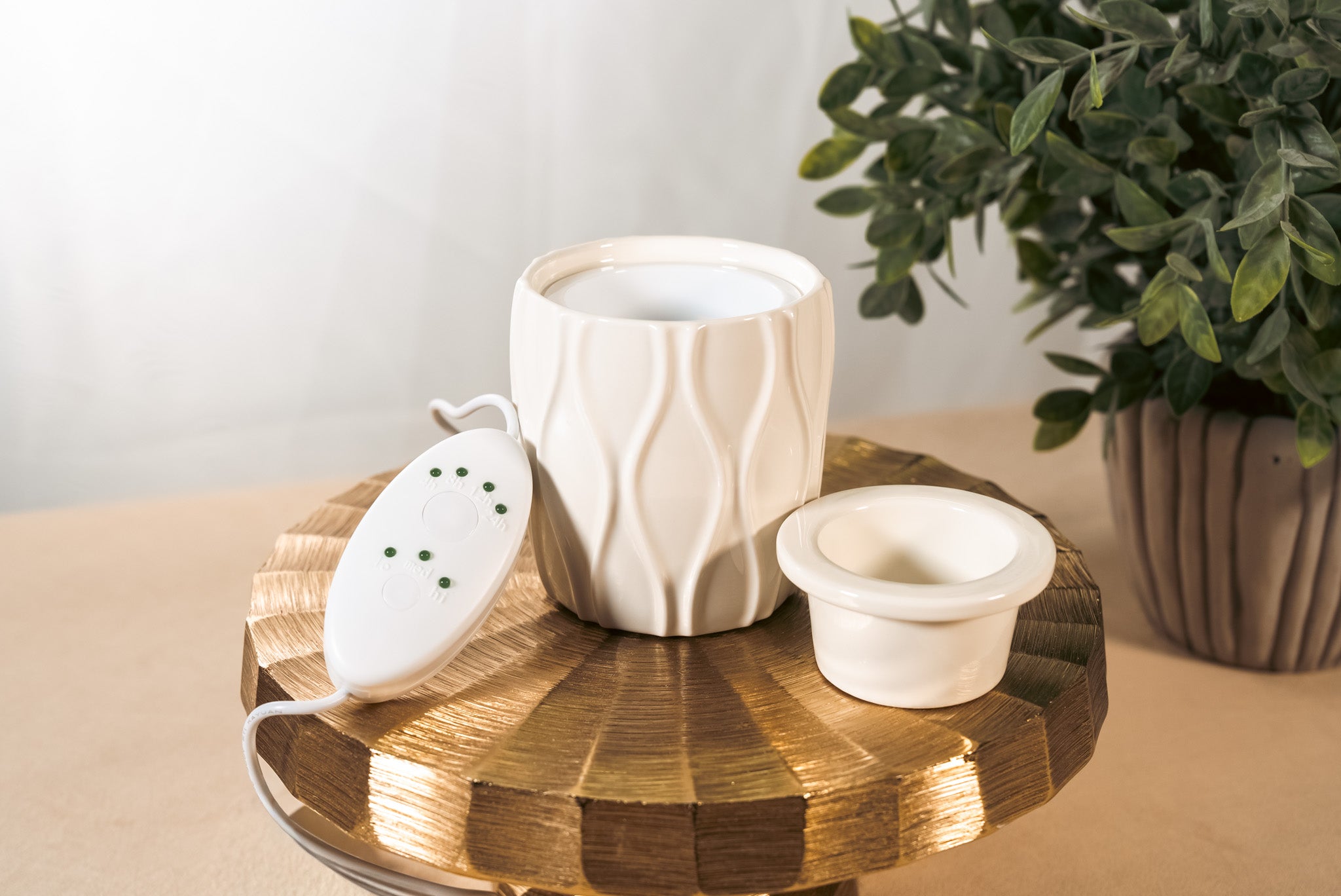 A textured, white fragrance warmer on a gold pedestal with a plant in the background