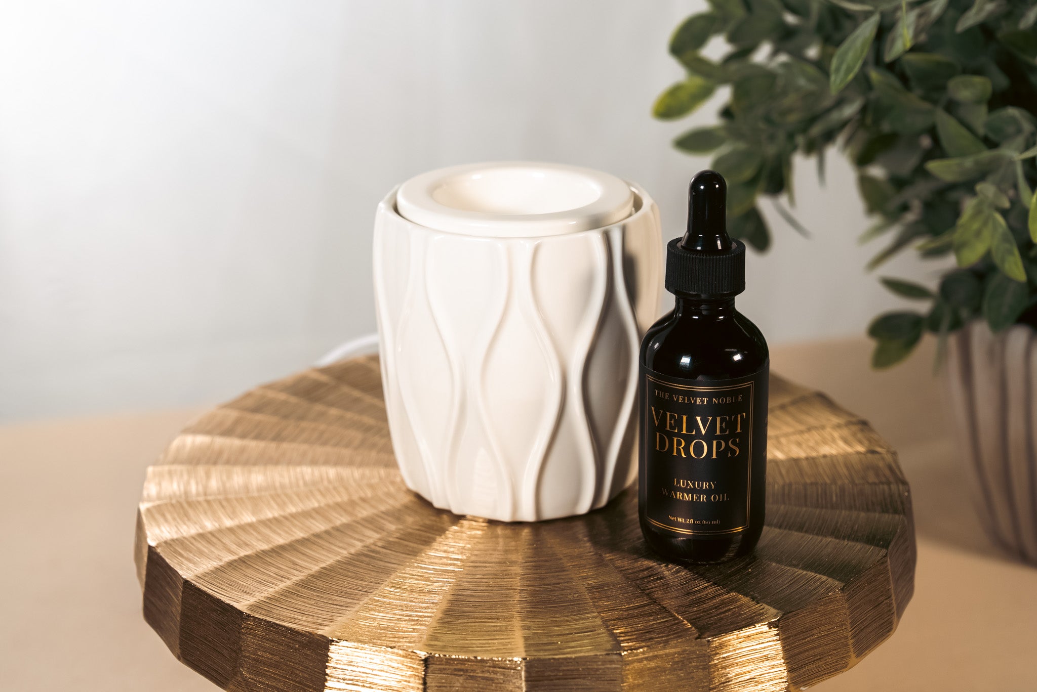 A textured, white fragrance warmer with a bottle of Velvet Drops on a gold pedestal with a plant in the background
