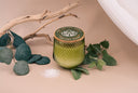 Green candle with wooden lid on a beige surface with leaves and a branch.
