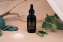 Bottle of 'Velvet Drops' oil with green leaves and branches on a beige background
