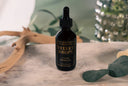 Black bottle of 'Velvet Drops' on a marble surface with greenery in the background