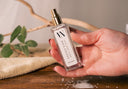 Hand holding a Mini Velvet Ease Air & Linen Mistr on a wooden surface with a blurred background