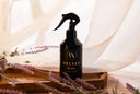 Black spray bottle labeled 'Velvet Home' on a wooden tray with lavender flowers, against a soft white curtain background.