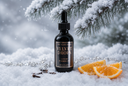 A bottle of Velvet Drops Luxury Warmer Oil stands in snow with evergreen branches overhead and orange slices nearby, creating a winter-themed product scene.