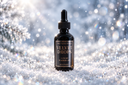 A dark amber glass dropper bottle labeled 'The Velvet Noble Velvet Drops Luxury Warmer Oil' standing upright in snow with a sparkling, bokeh winter background.