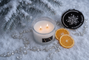 A glass candle with two lit wicks sits on a snowy surface beside two dried orange slices, jewels, and a black lid printed with a snowflake and the words 'DIAMOND FROST'. Flocked pine branches frame the top of the scene.