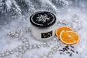 A glass jar candle with a black lid featuring a white snowflake and the text 'Diamond Frost' sits in a snowy winter scene. The candle is styled with jewels, orange slices, cloves, and pine branches.