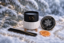 A black and white holiday-themed candle set styled in a snowy winter scene with pine branches, silver garland, dried orange slices, and a small black accessory piece. The candle jar is black with a gold monogram logo, the lid has a white snowflake and 'DIAMOND FROST' text, and the vessel is white ribbed ceramic.