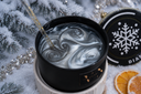 A black jar candle with a swirled white and gray wax design inside, set in a snowy holiday-themed scene with pine branches, silver tinsel, dried orange slices, and a snowflake-patterned ornament.
