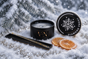 A black candle jar labeled 'VN' and 'DIAMOND FROST' sits in snow beside a matching lid with a snowflake emblem, dried orange slices, and a decorative tray, with snowy pine branches in the background.