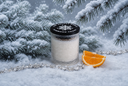 A glass jar candle with white wax and a black lid featuring a snowflake design, styled in a snowy winter scene with pine branches, silver tinsel, and orange slices.