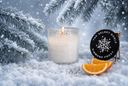 A lit white candle in a frosted glass holder sits in snow beside two orange slices and a round black label reading 'THE VELVET NOBLE DIAMOND FROST.' Snow-covered pine branches and falling snow create a winter scene.