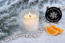 A lit candle in a textured clear glass holder sits in snowy faux snow with pine branches, silver tinsel, and orange slices. A round black product tag reading 'THE VELVET NOBLE DIAMOND FROST' is visible on the right.