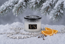 A glass candle jar labeled VN, styled in a snowy winter scene with pine branches overhead, jewels, cloves, and orange slices on white snow.