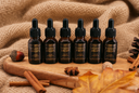 Six amber glass dropper bottles labeled VELVET DROPS arranged on a wooden board with autumn props including cinnamon sticks, a nut, star anise, and leaves, against a burlap fabric background.