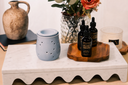 Three bottles of 'Velvet Drops' and a blue warmer on a wooden tray with decorative items including a vase and flowers.