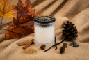 A candle in a textured clear glass jar sits on a beige knit blanket with autumn-themed props including brown leaves, pinecones, acorns, cinnamon sticks, vanilla beans, and a round black label reading 'THE VELVET NOBLE VANILLA HARVEST'.