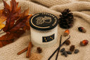 A 2-wick Vanilla Harvest candle on a woven blanket with autumn decor around it such as Vanilla beans, Cinnamon, Brown Sugar, Pinecones, acorns, and autumn leaves.