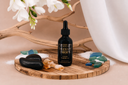 A 2 ounce bottle of Velvet Drops fragrance oil on a wooden tray with sea glass, sand, amber and florals on a beige background.