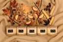 Five white candles in clear glass jars with black labels reading 'VN' are arranged on a beige textured fabric, surrounded by autumn-themed props including cinnamon sticks, apple slices, vanilla pods, and dried leaves.