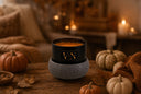 A black candle jar with a textured gray fragrance warmer sits on a rustic wooden table surrounded by small pumpkins, autumn leaves, and warm candlelight in the background.