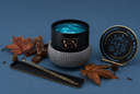 A scented wickless candle from The Velvet Noble, featuring a black jar with blue wax on a gray textured warmer, its matching lid on a small stand, styled with autumn leaves, cinnamon sticks, and pinecones on a blue background.