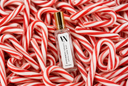 A clear rectangular perfume bottle with a gold cap and white label reading 'VN Peppermint Legacy' lies centered on a dense bed of red-and-white striped candy canes.