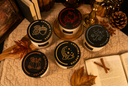 Five jars with black lids arranged on a quilted cream blanket with autumn-themed props including a book, cinnamon sticks, pinecone, and dried leaves. The lids feature different illustrated designs.