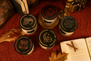 Five jars with black lids arranged on a quilted red blanket with autumn-themed props including a book, cinnamon sticks, pinecone, and dried leaves. The lids feature different illustrated designs.