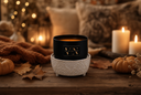 A black candle jar with gold lettering sits in a textured white ceramic holder on a rustic wooden table, surrounded by autumn decor including pumpkins, dried leaves, a knitted blanket, and lit candles in the background.