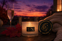 A lit scented candle in a textured glass jar sits on a wooden table at sunset, with a champagne flute, red leaves, a knitted blanket, and a round black lid beside it. The candle label reads VN, and the lid shows a crescent moon with the words Golden Twilight.