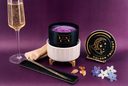 A luxury wickless candle from VN VELVET NOBLE featuring a black and white candle in a ribbed ceramic warmer, its gold lid, a gold stirring stick, and decorative flowers on a purple background.