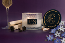 A lit scented candle in a textured clear glass jar sits on a purple surface, with a matching round black lid placed to the right. The label reads The Velvet Noble and GOLDEN TWILIGHT.