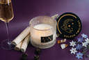 A lit wax candle in a textured clear glass jar sits on a purple surface with a black round tin lid beside it, a champagne flute, wooden sticks, crystals, and small white and blue flowers. The lid shows the name Golden Twilight.