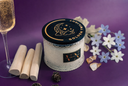 A glass jar candle with a black lid featuring a gold crescent moon and stars design and the words 'GOLDEN TWILIGHT' is styled on a purple background with wooden sticks, a glass of sparkling liquid, and small white and blue flowers.
