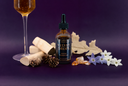 A bottle of Velvet Drops Luxury Warmer Oil centered on a dark purple background, styled with a wine glass, dried leaves, pinecones, bark pieces, and small white flowers.