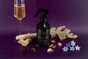 A black spray bottle labeled 'VELVET home' stands on a purple surface with decorative props including a filled champagne flute, rolled wood pieces, pinecones, amber stones, and small blue and white flowers.