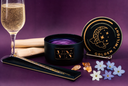 A black candle labeled 'VIN The Velvet Noble' with a purple shimmer wax surface, shown beside its matching lid on a stand. The scene is styled with a filled champagne flute, a wooden stick, crystals, and small white and lavender flowers on a purple background.