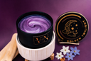 A black candle jar with a purple and lavender swirl wax inside, displayed next to its black lid featuring a gold crescent moon and stars design. The candle is set in a white ribbed ceramic warmer on a small wooden stand, with decorative flowers and a wooden spoon on a dark purple background.