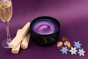 A black candle jar with a purple and lavender swirled shimmering wax inside, styled on a purple background with a glass of sparkling wine, wooden sticks, amber crystals, and small blue and white flowers.