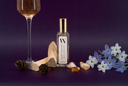A styled flat-lay featuring a bottle of Golden Twilight Mini Air & Linen Mist with a gold cap, surrounded by a glass of sparkling wine, wooden rollers, pinecones, raw amber, blue and white flowers, and a tarot card on a deep purple background.