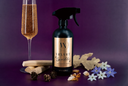 A dark spray bottle labeled 'Velvet Sparkle' stands on a purple surface with a filled champagne flute, dried leaves, pinecones, and small flowers around it.