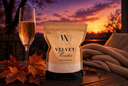A stand-up pouch of Velvet Powder luxury freshener stands on a wooden table outdoors at sunset, with a filled champagne flute, autumn leaves, and a knitted blanket nearby. The package is cream and black with visible white powder at the bottom.