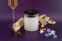 A textured glass jar candle with a black lid featuring gold lettering, styled on a purple background with natural elements like pinecones, wood slices, dried leaves, and small flowers.