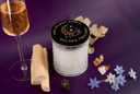 A glass jar candle with a black lid featuring gold lettering and an illustration, styled on a purple background with a glass of sparkling golden liquid, wooden sticks, crystals, and small blue and white flowers.