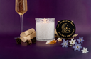 A lit white wax candle in a clear textured glass holder sits on a purple surface, styled with a champagne flute, rolled parchment, pinecones, crystals, and small white flowers. A round black coaster reads 'The Velvet Noble Golden Twilight'.