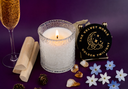 A lit white candle in a textured clear glass holder on a purple surface, styled with a glass of sparkling golden liquid, wooden sticks, crystals, blue and white flowers, and a round black coaster printed with 'THE VELVET NOBLE GOLDEN TWILIGHT'.